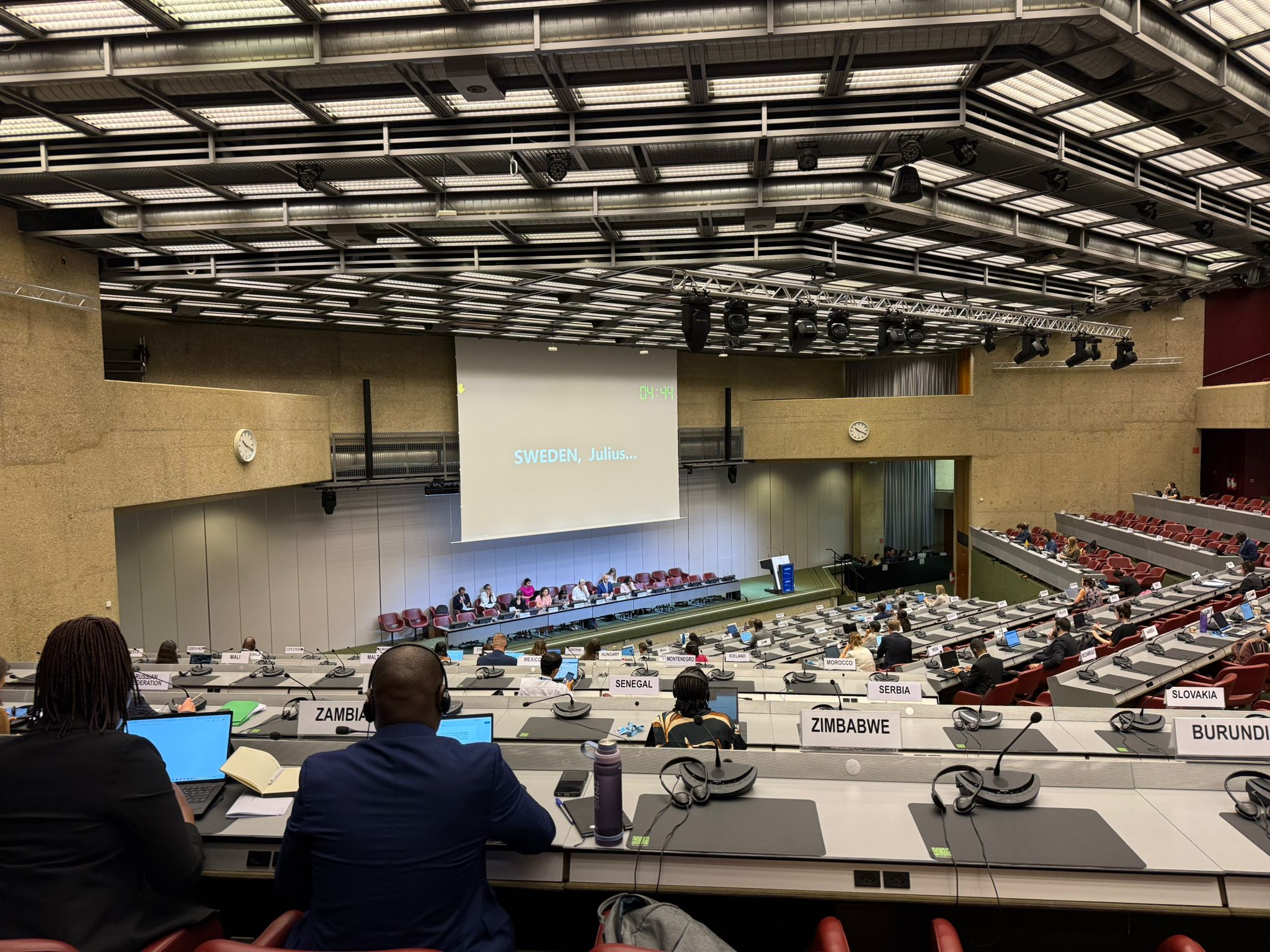 A panel discussion in a conference hall with delegates seated at desks.